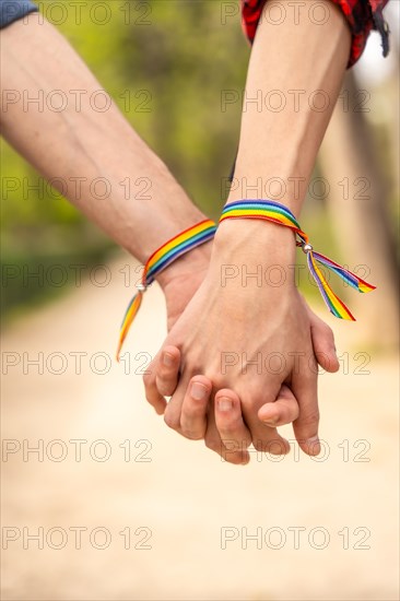 Vertical close-up photo of two unrecognizable gay people with lgbt bracelet holding hands in a park