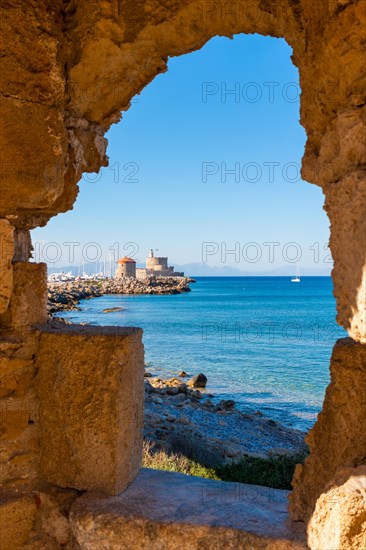 View of the bay of Rhodes through a window in a rock wall. In the background, the sea and the windmill in the Mandraka port