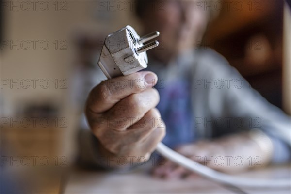 Senior citizen holding a power cable with plug in her hand at home, symbolising energy costs and poverty, Cologne, North Rhine-Westphalia, Germany, Europe