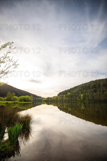 A lake in a landscape shot. A sunset and the natural surroundings are reflected in the water of the reservoir. Marbach reservoir, Odenwald, Hesse