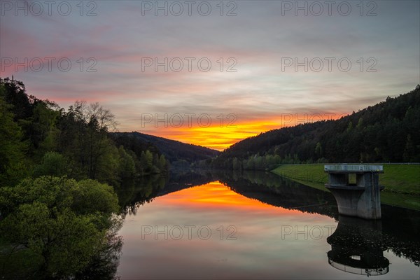 A lake in a landscape shot. A sunset and the natural surroundings are reflected in the water of the reservoir. Marbach reservoir, Odenwald, Hesse