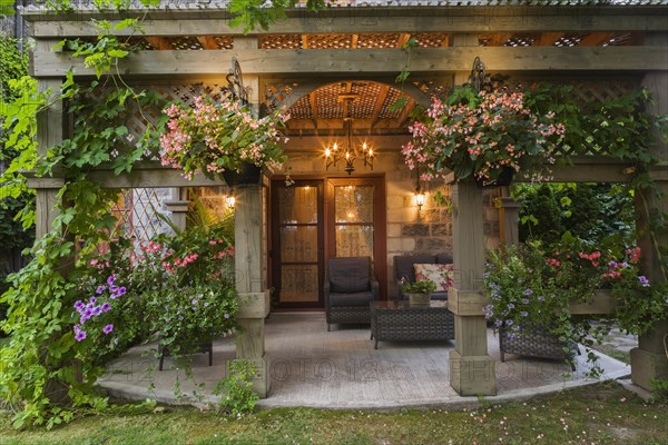 Rear of Renaissance grey stone and mortar castle style home with concrete patio and lattice covered wooden gazebo decorated with climbing Vitis, Vines, purple Petunias and pink Begonia flowers in hanging baskets in summer at dusk, Quebec, Canada, North America