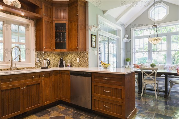 Kitchen with cherry wood cabinets, earth tone slate flooring and wooden breakfast table with sitting bench and rustic chairs inside a contemporary cottage style home, Quebec, Canada, North America