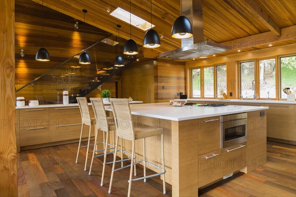 Bamboo wood island with white quartz countertops and high wicker chairs, buffet in kitchen with Ipe wood floor plus black industrial style pendant lighting fixtures inside luxurious stained cedar and timber wood home with panoramic windows, Quebec, Canada, North America