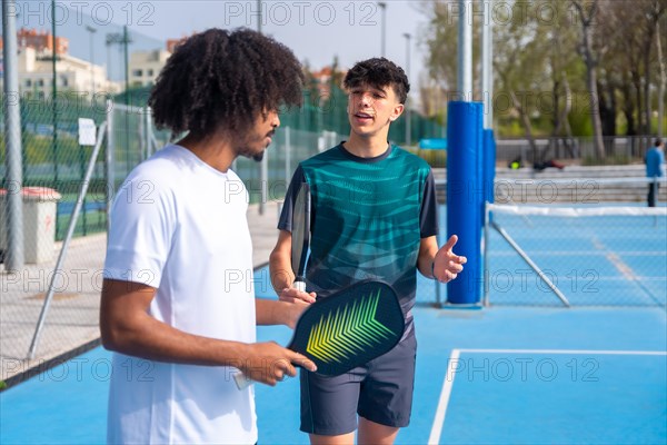 Caucasian young male instructor talking to a young african man playing pickleball in an outdoor court during sunny day