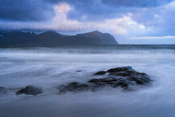 On the beach at Vikten. The sea washes around some rocks. Mountains in the background. At night at the time of the midnight sun, cloudy sky. Early summer. Long exposure. Vikten, Flakstadoya, Lofoten, Norway, Europe