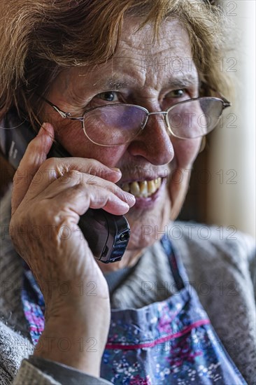 Laughing senior citizen with smock talking on the phone at home in her living room, Cologne, North Rhine-Westphalia, Germany, Europe