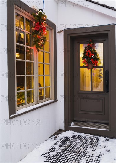 Window and brown front entrance door with illuminated Christmas lights and decorations on an old circa 1886 white with beige and brown trim Canadiana cottage style home at dusk in winter, Quebec, Canada, North America