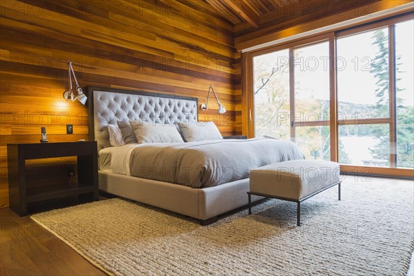 King size bed with grey upholstered panel headboard, ottoman and wooden end tables in master bedroom with grey nuanced rug and Ipe wood floor inside luxurious stained cedar and timber wood home with panoramic windows, Quebec, Canada, North America