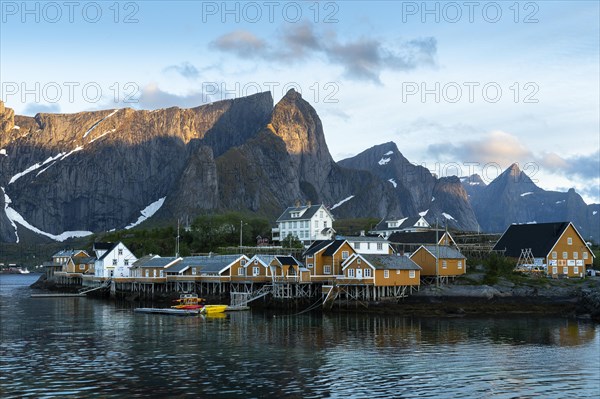 The village of Sakrisoy with its typical yellow or ochre-coloured wooden houses on wooden stilts (rorbuer) by the sea. At night at the time of the midnight sun in good weather. Blue sky, few clouds. Sunlight on the mountain peaks. Early summer. Sakrisoy, Moskenesoya, Lofoten, Norway, Europe