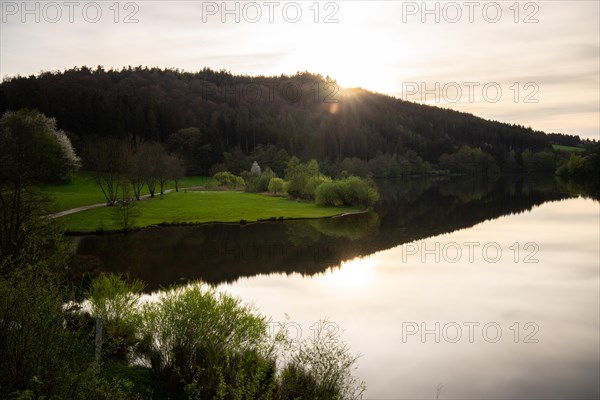 A lake in a landscape shot. A sunset and the natural surroundings are reflected in the water of the reservoir. Marbach reservoir, Odenwald, Hesse