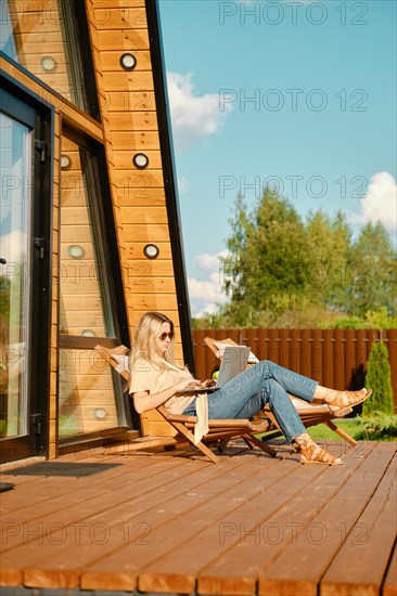 Woman typing on laptop while sitting in a folding wooden sun lounger on the terrace of a-frame cabin