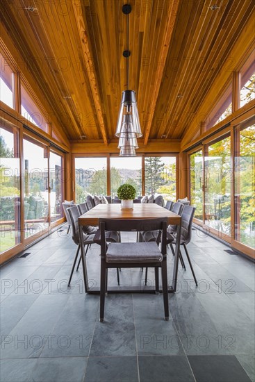 Four season gazebo and industrial style black with clear glass pendant lighting fixtures over wood and black metal dining table with grey upholstered chairs and heated slate floor inside luxurious stained cedar and timber wood home with panoramic windows, Quebec, Canada, North America