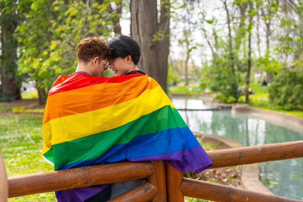 Rear view of a romantic scene of a multi-ethnic gay couple in love wrapped in lgbt flag embraced in a park