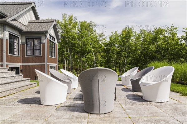 White and grey cloth covered sitting chairs on backyard patio and rear view of contemporary natural stone and brown stained wood and cedar shingles clad luxurious bungalow style home in summer, Quebec, Canada, North America