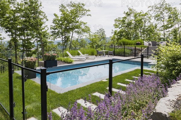 Two white long chairs on edge of in-ground swimming pool enclosed by clear glass and black metal fence in residential backyard in summer, Quebec, Canada, North America