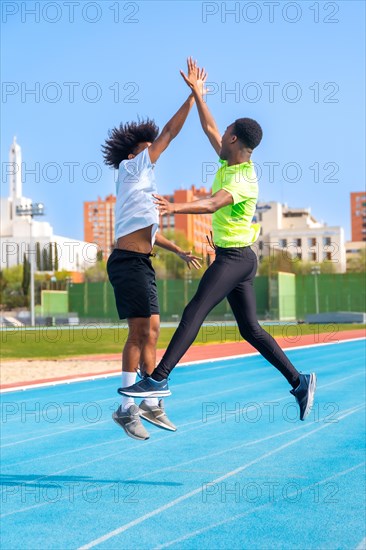 Vertical photo of two african american young friends jumping celebrating and clasping hands in a running track