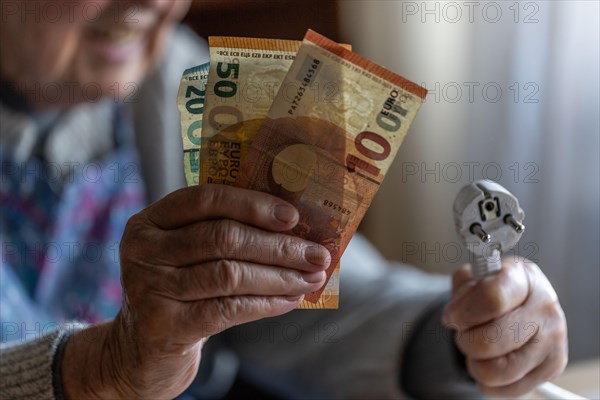 Senior citizen holding money and a power cable with plug in her hand at home, symbolising energy costs and poverty, Cologne, North Rhine-Westphalia, Germany, Europe