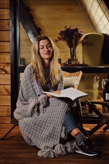 A woman wrapped in a plaid sits on the terrace in the evening and reads a book