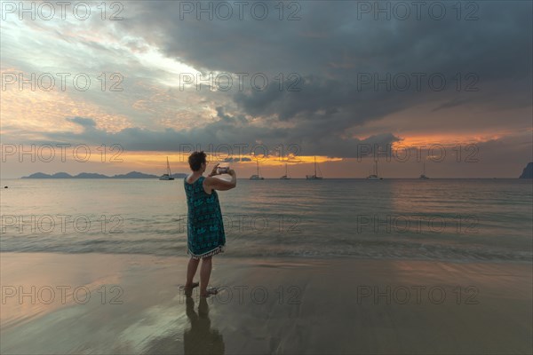 Woman, 60-65, taking a photo of the sunset with her smartphone, Koh Mook Island, Andaman Sea, Thailand, Southeast Asia, Asia