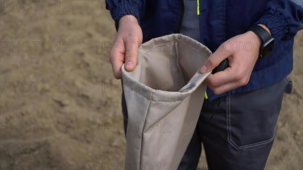 A child in a blue jacket and jeans stands on the sand and holds a bag of clothes