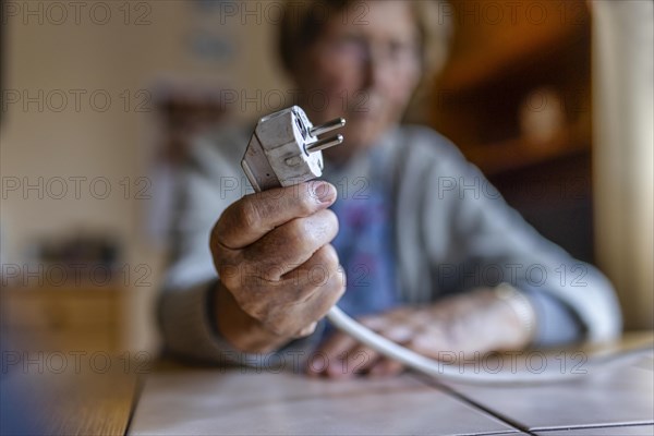 Senior citizen holding a power cable with plug in her hand at home, symbolising energy costs and poverty, Cologne, North Rhine-Westphalia, Germany, Europe