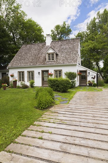 Old circa 1886 white with beige and brown trim Canadiana cottage style home facade and wood plank driveway in summer, Quebec, Canada, North America