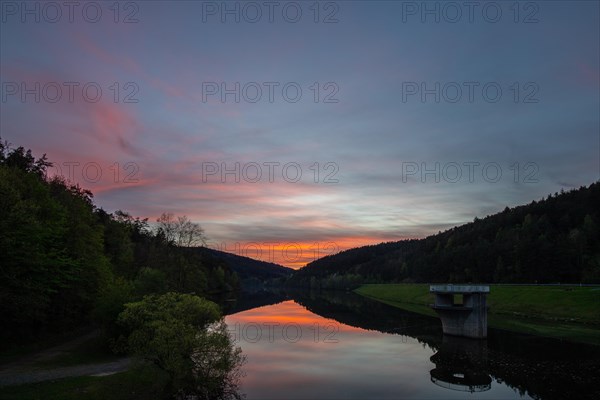 A lake in a landscape shot. A sunset and the natural surroundings are reflected in the water of the reservoir. Marbach reservoir, Odenwald, Hesse