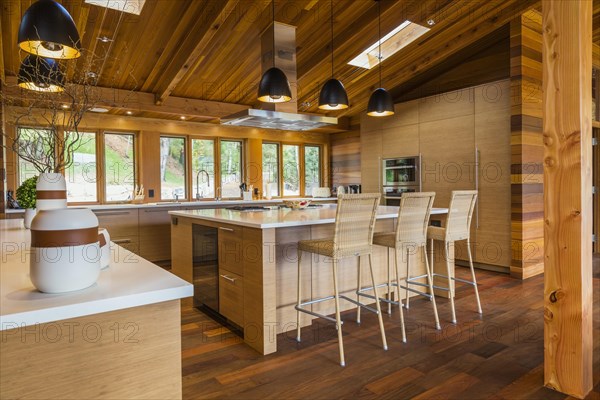 Bamboo wood buffet and island with white quartz countertops and high wicker chairs, cabinets in kitchen with Ipe wood floor plus black industrial style pendant lighting fixtures inside luxurious stained cedar and timber wood home with panoramic windows, Quebec, Canada, North America