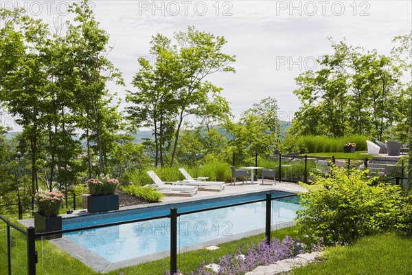 Two white long chairs on edge of in-ground swimming pool enclosed by clear glass and black metal fence in residential backyard in summer, Quebec, Canada, North America