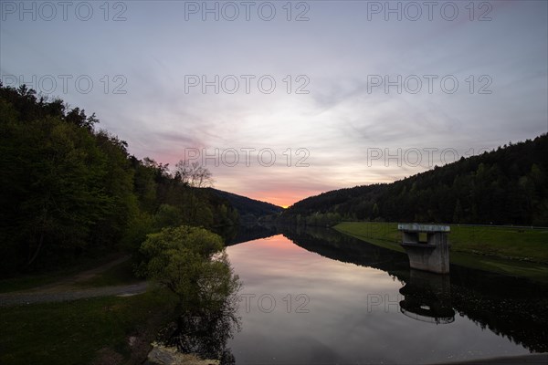 A lake in a landscape shot. A sunset and the natural surroundings are reflected in the water of the reservoir. Marbach reservoir, Odenwald, Hesse