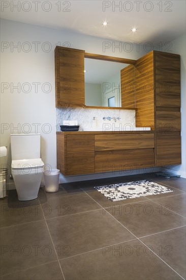 White high-back flush toilet and American walnut wood vanity with white rectangular sink and ceramic tile flooring in main bathroom on upstairs floor inside modern cube style home, Quebec, Canada, North America