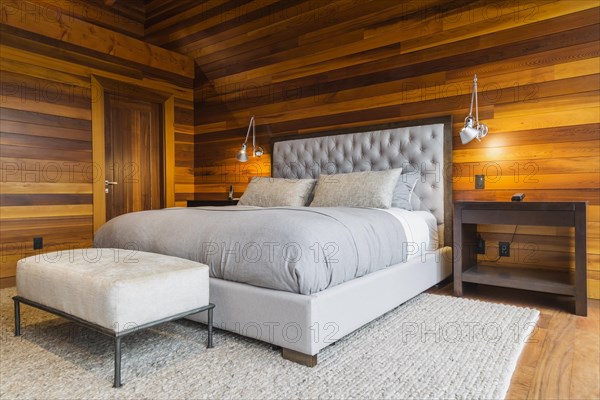 King size bed with grey upholstered panel headboard, ottoman and wooden end tables in master bedroom with grey nuanced rug and Ipe wood floor inside luxurious stained cedar and timber wood home, Quebec, Canada, North America