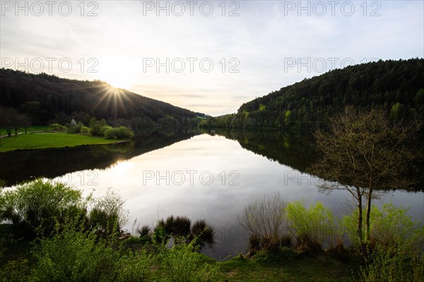 A lake in a landscape shot. A sunset and the natural surroundings are reflected in the water of the reservoir. Marbach reservoir, Odenwald, Hesse