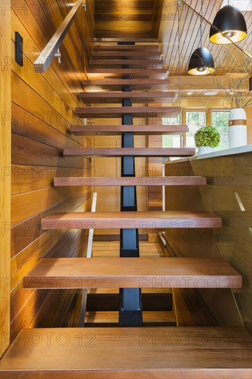 Looking up illuminated wooden staircase with opened steps and clear glass railing leading to upstairs floor inside luxurious stained cedar and timber wood home, Quebec, Canada, North America