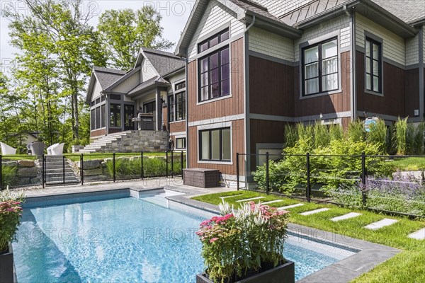 In-ground swimming pool and rear view of contemporary natural stone and brown stained wood and cedar shingles clad luxurious bungalow style home in summer, Quebec, Canada, North America