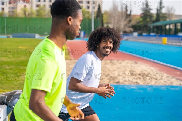 Side view of two smiling african young friends stretching and warming up in a running track