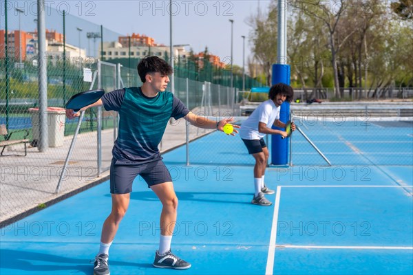 Caucasian young man serving ball playing pickleball with african american young male partner in an outdoor court