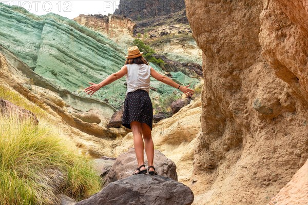A tourist woman with a hat and open arms at the Azulejos de Veneguera or Rainbow Rocks in Mogan, Gran Canaria