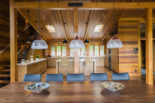 Industrial style copper with frosted glass pendant lighting fixtures over wooden dining table with black leather chairs plus kitchen with bamboo wood island and cabinets in background inside luxurious stained cedar and timber wood home with panoramic windows, Quebec, Canada, North America