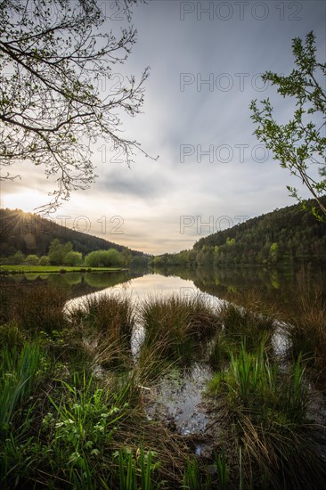 A lake in a landscape shot. A sunset and the natural surroundings are reflected in the water of the reservoir. Marbach reservoir, Odenwald, Hesse