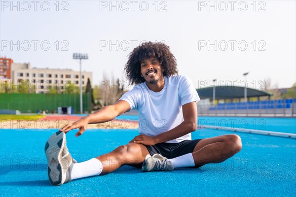 Frontal portrait of an African smiling man with curly hair and sports clothes stretching in an outdoor running track