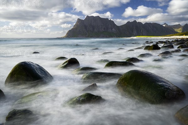 Seascape on the beach at Uttakleiv (Utakleiv) . Rocks in the sea. Round stones surrounded by water. Mount Hogskolmen in the background. Sun and clouds. Early summer. Long exposure. Uttakleiv, Vestvagoya, Lofoten, Norway, Europe