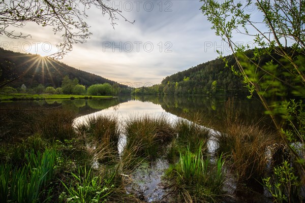 A lake in a landscape shot. A sunset and the natural surroundings are reflected in the water of the reservoir. Marbach reservoir, Odenwald, Hesse