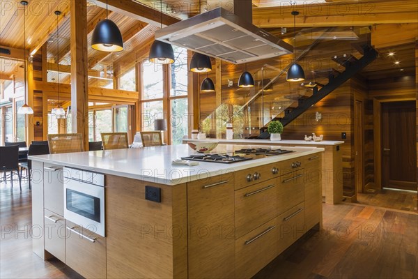 Bamboo wood island with white quartz countertops and high wicker chairs, buffet in kitchen with Ipe wood floor plus black industrial style pendant lighting fixtures inside luxurious stained cedar wood and timber home with panoramic windows, Quebec, Canada, North America