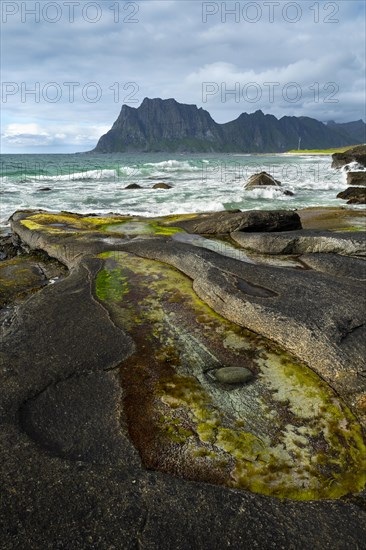 Seascape on the beach at Uttakleiv (Utakleiv), with rocks and water-filled rocky outcrops in the foreground. Mount Hogskolmen in the background. Sun and clouds. Early summer. Uttakleiv, Vestvagoya, Lofoten, Norway, Europe