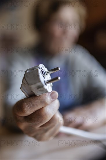 Senior citizen holding a power cable with plug in her hand at home, symbolising energy costs and poverty, Cologne, North Rhine-Westphalia, Germany, Europe