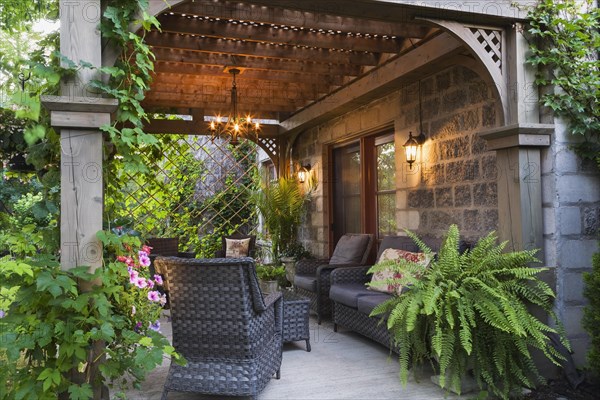 Rear of Renaissance grey stone and mortar castle style home with concrete patio and lattice covered wooden gazebo decorated with climbing Vitis, Vines, purple Petunias and Adiantum palmatum, Maidenhair Ferns in planter in summer at dusk, Quebec, Canada, North America
