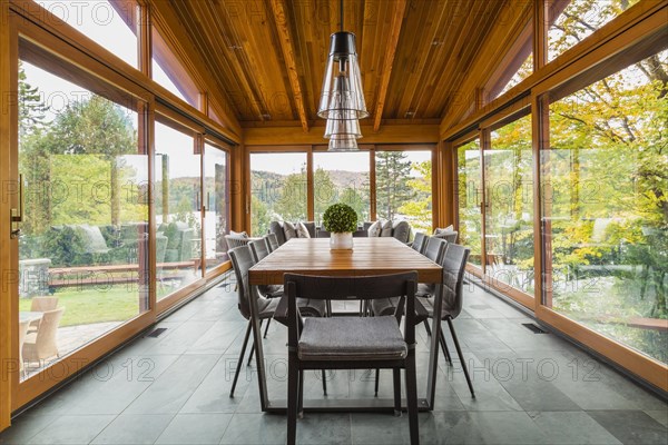 Four season gazebo and industrial style black with clear glass pendant lighting fixtures over wood and black metal dining table with grey upholstered chairs and heated slate floor inside luxurious stained cedar and timber wood home with panoramic windows, Quebec, Canada, North America