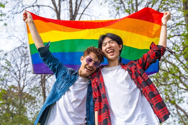 Low angle view portrait of a happy gay couple laughing while raising lgbt rainbow flag outdoors in a park
